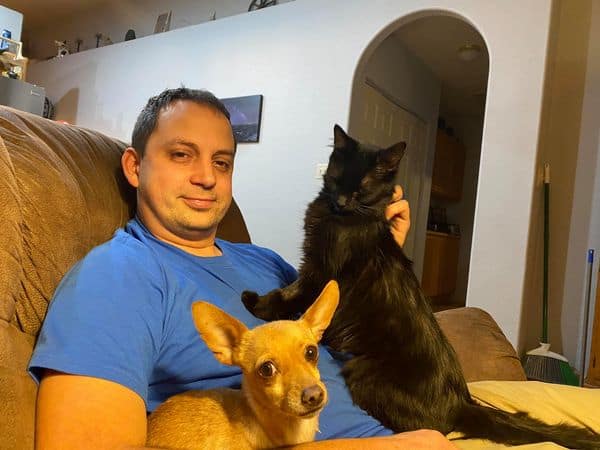 A man sitting on a couch with his brown dog and black cat