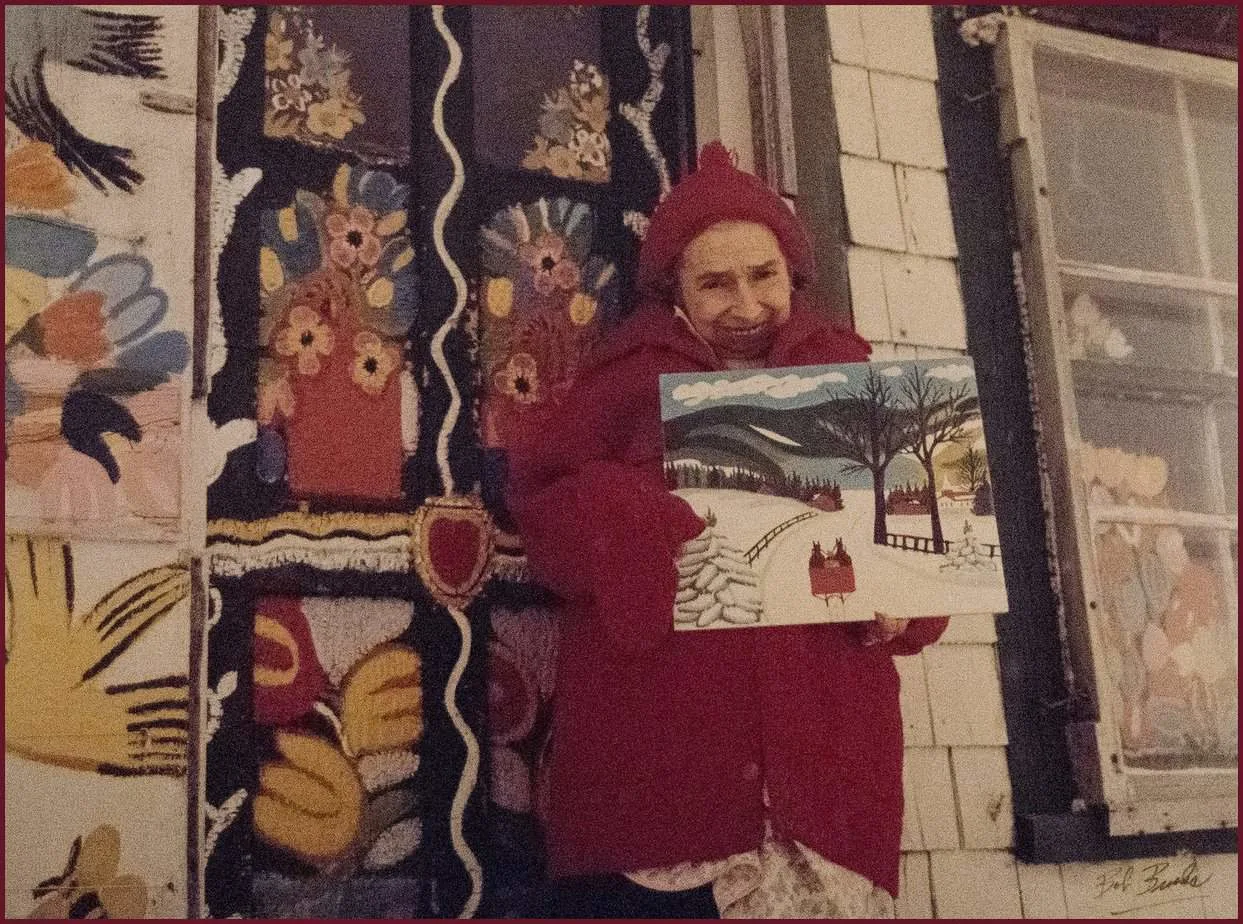 Maud Lewis holding up a painting of hers in front of her house