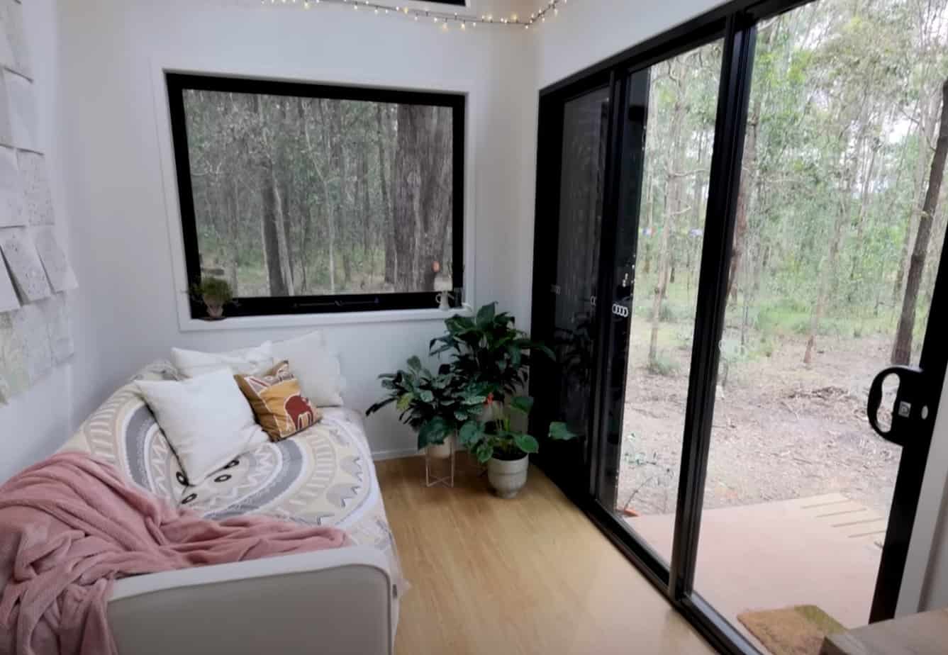 The tiny house's living room has nice couch and amazing view of the woods from its huge windows.