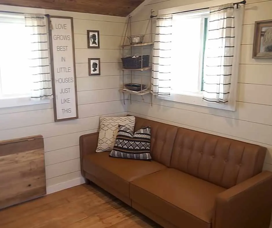 Krebs' tiny home's living area with large sofa, windows and a wall display that says "love grows best in little houses just like this"